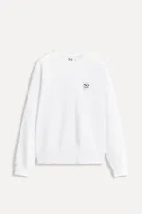 Sudadera blanca de cuello redondo y manga larga, confeccionada en algodón. Tiene un parche con el logo del 50 aniversario en el pecho y terminaciones en rib a tono.