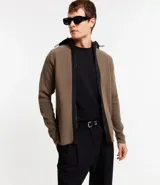 Cardigan de punto color marrón topo, con capucha negra contrastante y cierre frontal de cremallera negra. Presenta bolsillos laterales.