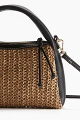 Cartera pequeña con cuerpo de paja trenzada color marrón claro y laterales de cuero sintético negro. Tiene asa corta de mano y correa larga extraíble para usar cruzada.