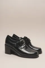 Zapato de cuero vacuno charol negro, con punta redondeada y detalle de perforado en la capellada. Tiene una tira con hebilla metálica y un taco cuadrado de 4 cm.