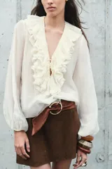Blusa color crudo confeccionada en hilatura con mezcla de lyocell. Escote pico ajustable con cordones, manga larga con volumen acabada en elástico y detalle de volantes en el mismo tejido.