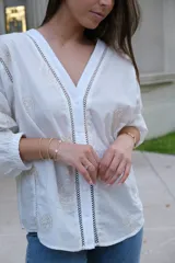 Blusa blanca de manga larga con elástico en puños, escote en pico y cierre frontal con botones. Detalles de bordados con hilado en tonalidad camel y acabado con entredoses a tono.