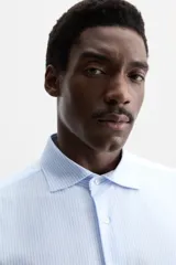 Camisa celeste a rayas verticales blancas, de corte recto, con cuello italiano abierto, manga larga con puños abotonados y cierre frontal con botones.