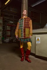 Chaqueta con cuerpo de lana a cuadros en tonos marrón, verde y amarillo. Mangas y cuello de cuero agamuzado color camel. Cierre frontal con cremallera. Falda corta a cuadros escoceses en tonos verde, rojo y azul. Medias amarillas y botas de cuero marrón. 