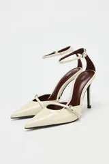 Zapato de tacón alto fino, color beige acharolado, con punta fina y tira con hebilla en la parte delantera y tobillo.