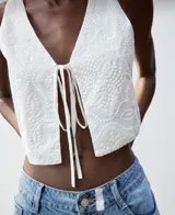 Chaleco crop top blanco con bordado floral y lazos frontales.