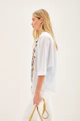 Camisa blanca de voile con rayas finas, corte amplio, cartera con botones y bordado de flores en el frente.