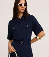 Camisa color beige de tela airflow, con cuello camisero, mangas cortas con detalle de pliegue, bolsillos con solapa en el pecho y cierre frontal con botones metalizados.