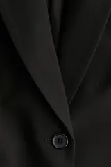 Blazer negro de corte entallado con solapas de muesca, cierre de un botón y hombreras. Presenta bolsillos delanteros ribeteados y abertura en la espalda.