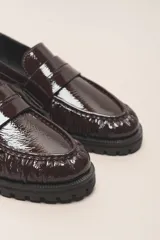 Mocasín negro de cuero vacuno con acabado charol, suela de goma y forro y plantilla de cuero vacuno.