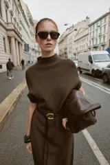 Vestido negro de tejido de punto, corte midi y silueta holgada, con cuello alto acanalado y mangas cortas tipo murciélago. Se ajusta a la cintura con un cinturón fino de efecto piel negro con hebilla dorada en forma de C.