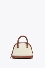 Bolso de mano color crema con detalles en cuero marrón. Tiene forma de domo, cierre superior con cremallera, bolsillo interno con cremallera y correa bandolera extraíble.