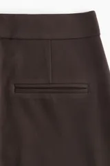 Pantalón de sarga color marrón oscuro, de tiro alto con elástico oculto en la cintura, cierre y pretina prolongada con botón y corchetes ocultos. Tiene piernas con corte para bota, bolsillos al bies, bolsillos de atrás ribeteados y pliegues adelante y atrás.