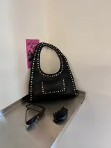 Cartera negra tipo hobo de ecocuero con tachas plateadas y bolsillo frontal.