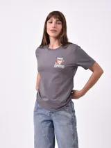 Remera blanca de algodón con cuello redondo y manga corta. Presenta un pequeño bordado en el pecho izquierdo con una figura que parece ser una persona acostada en una tabla de surf o flotador.