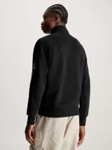 Buzo negro de felpa de algodón orgánico con cuello alto con cremallera, puños y bajo de canalé, bolsillo canguro y parche con logo en la manga.