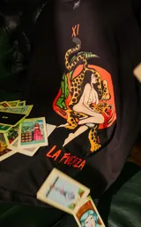 Remera negra de manga corta con cuello redondo y calce oversize. Presenta una estampa en la espalda inspirada en el tarot, con la carta "La Fuerza" que muestra a una mujer desnuda sobre un leopardo, rodeada de vegetación y un sol rojo.