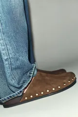 Zueco destalonado de piel color marrón con acabado en punta redonda. Presenta detalle de tachas doradas en los laterales y pespunte decorativo en la parte delantera. Posee una suela plana con un tacón mínimo de 0,5 cm.