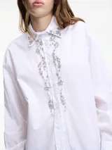 Camisa blanca de algodón con cuello camisero, cierre frontal con botones y apliques de pedrería en cuello y cartera de botones.