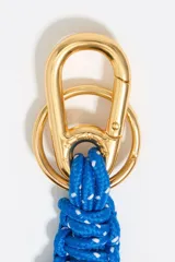 Llavero con mosquetón metálico dorado, cordón trenzado azul con motas blancas y dije en forma de corazón naranja con centro dorado y el nombre de la marca grabado.