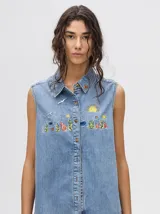 Camisa sin mangas de denim ligero, color celeste lavado, con cuello camisero y botones frontales. Presenta bordados temáticos de playa/mar en el pecho, incluyendo sol, olas, gaviotas, peces y corales/vegetación marina.