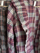 Camisa de tela escocesa liviana, color principal bordó con cuadros en tonos beige, gris y naranja. Presenta volados en el frente y mangas abullonadas.