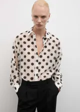 Camisa color crema con estampado de lunares negros, de corte recto, manga larga con puños abotonados, cuello camisero y cierre frontal oculto con tapeta.