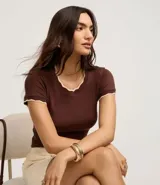 Blusa de algodón con textura acanalada en color blanco, de manga corta y cuello redondo. Presenta un acabado ondulado tipo festón con borde contrastante en color negro tanto en el cuello como en los puños de las mangas.