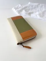 Billetera grande con cierre metálico, confeccionada en cuero con diseño de bloques de color: verde oliva texturizado, marrón claro liso y blanco/crema texturizado. Posee un monedero interno y un tirador de cuero marrón claro.