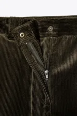 Pantalón de pana color verde oscuro, de tiro alto y corte ancho. Tiene bolsillos delanteros y trabillas en la cintura. Cierre frontal con cremallera y botón metálico.