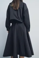 Falda midi negra de gabardina, tiro medio, cintura con trabillas, corte evasé con volumen y cierre frontal con cremallera y botón.