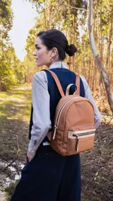 Mochila marrón de nylon con detalles en símil cuero en el bolsillo frontal. Tiene asa de mano, correas regulables y un bolsillo trasero vertical. El interior es espacioso, con dos bolsillos con cierre y uno abierto con división.