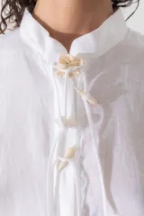 Túnica blanca de lino, corte recto, cuello mao, manga corta abullonada y cierre frontal con lazos.
