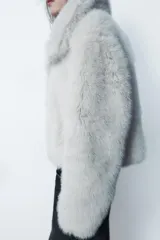 Abrigo corto color blanco de efecto piel, con cuello solapa y manga larga. Tiene bolsillos laterales ocultos en la costura, forro interior combinado y cierre frontal con ganchos metálicos forrados.