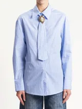 Camisa de manga larga con cuello camisero, confeccionada en tela de rayas diplomáticas celestes y blancas. Incluye un corbatín a juego sujeto con un broche tipo camafeo.