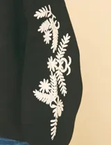 Suéter de punto negro con cuello redondo y bordados florales en hilo dorado en ambas mangas.