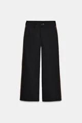Pantalón negro con finas rayas verticales blancas, de tiro medio y corte ancho. Presenta bolsillos delanteros y traseros, cintura doble tipo bóxer con botones y cierre frontal con cremallera y ganchos metálicos. Detalle de franja marrón en los laterales.