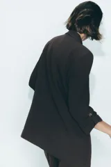 Blazer color marrón oscuro, abierta, con cuello solapa y hombreras. Manga tres cuartos con trabilla y botón dorado. Bolsillos delanteros con solapa.