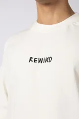 Remera color crema de corte oversize con la palabra "Rewind" bordada en el frente.