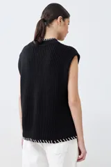 Chaleco negro de tejido de punto con cuello alto, cierre parcial con botones y detalle de pespunte blanco en el ruedo y el cuello. Sin mangas.