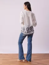 Blusa blanca de gasa bambula con cuello a la base, escote en V y pliegues. Tiene apertura central con botones, mangas ¾ con terminación fruncida y ruedo con detalles bordados en negro.