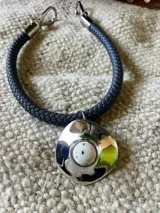 Collar con cordón trenzado de cuero sintético color negro, con dije circular plateado con centro de piedra blanca con pintitas negras. Cierre con mosquetón plateado.