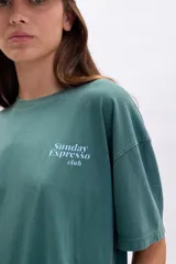Remera verde con corte oversize y estampa en la espalda con la frase "Sunday Expresso Club" y un dibujo de un gato dentro de una taza de café.