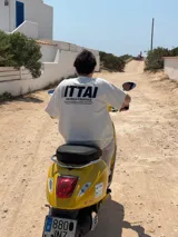 Remera color crema de corte recto y mangas cortas, con estampado en la espalda con el logo "ITTAI" en negro con líneas rojas, azules y blancas, y la leyenda "AN IBIZA PRODUCE. Ittai Stars Department Gallery - Ibiza Edition. If you're in Ibiza, 112 is not an option.".