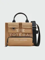 Bolso tote mediano de lona color beige con detalles en negro. Tiene doble asa de mano de cuero y correa bandolera ajustable y extraíble. Cuenta con dos bolsillos frontales con solapa y un bolsillo frontal con cierre. Estampado frontal con la leyenda "THE TOTE BAG" y el logo "MARC JACOBS".