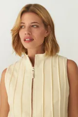 Chaleco color crema con cuello alto y cierre frontal con cremallera. Diseño con alforzas verticales en la parte superior delantera y trasera.