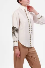 Camisa blanca de poplin de algodón con cuello clásico, cierre frontal con botones y mangas largas con puños abotonados. Presenta detalle de estampado de leopardo en blanco y negro en los puños y detalle de triángulos bordados en beige a lo largo del frente.