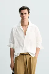 Camisa blanca de corte relajado confeccionada en tejido de algodón y lino. Tiene cuello bowling, manga larga con puños abotonados, jaretas en el delantero, bolsillos de parche en pecho y cadera, bajo con aberturas laterales y cierre frontal con botones.