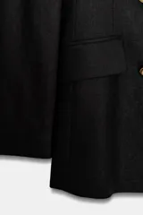 Blazer cruzada de color negro, confeccionada en tejido con lino. Presenta cuello solapa con muescas, hombreras, manga larga, falsos bolsillos con solapa y cierre frontal cruzado con botones metálicos dorados.