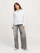 Camisa blanca de corte oversize con cuello clásico, cierre frontal con botones y mangas largas con puños abotonados. Presenta pequeños brillantes plateados distribuidos por toda la prenda.